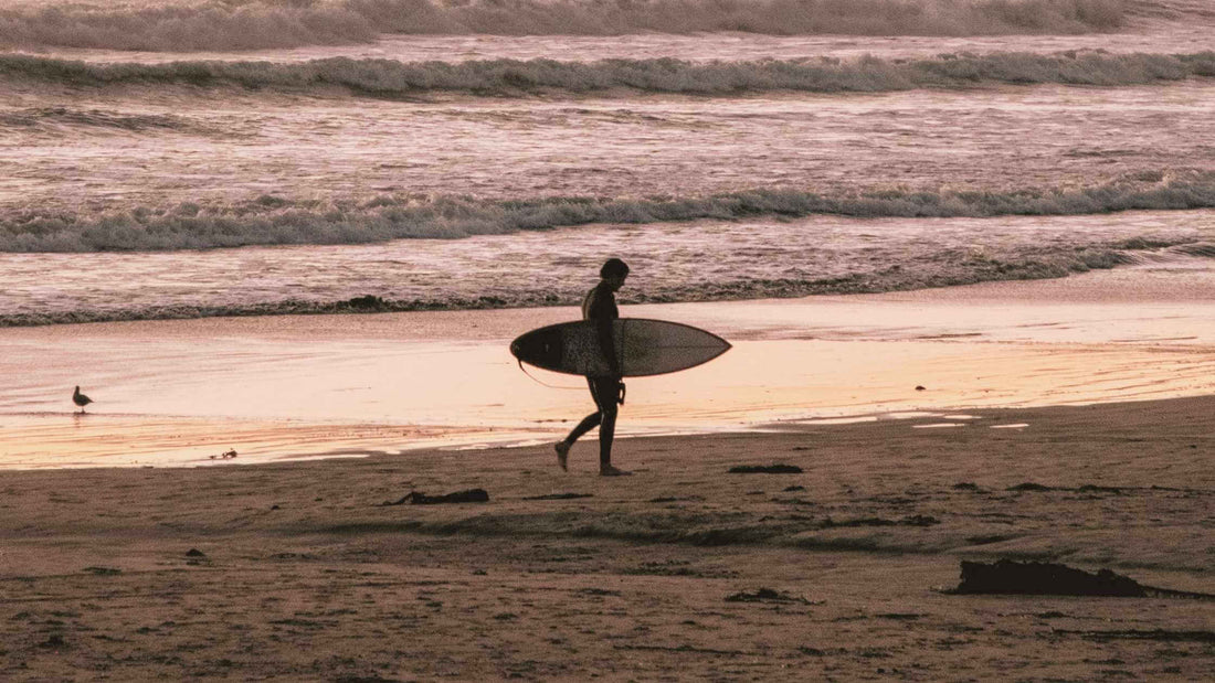 Surfer walking the beaches at dusk - Read about surfing and surf wear at Brag Vintage